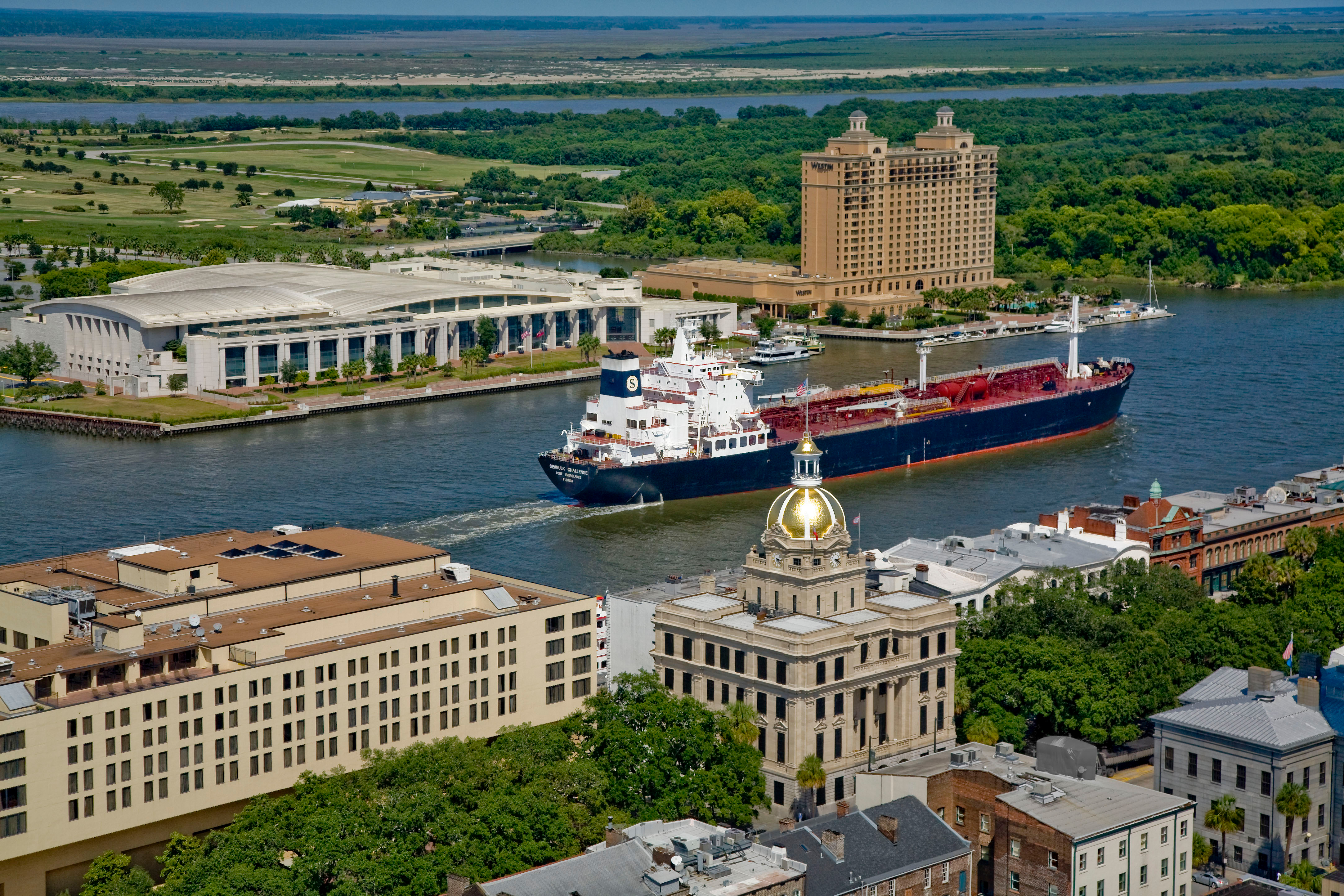 An aerial view of Savannah that includes the Riverfront District, Convention Center, City Hall, and a ship cruising down the Savannah River.